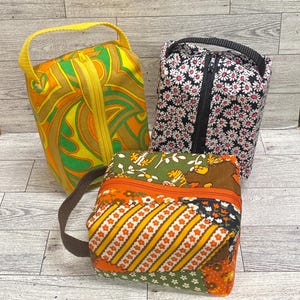 May include: Three small zippered pouches with different patterns. The first pouch is yellow, green, and orange with a swirling pattern. The second pouch is black with white daisy flowers. The third pouch is a patchwork of orange, green, and brown with a variety of patterns.