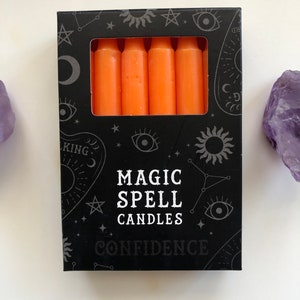 Könnte beinhalten: Eine schwarze Box mit einem silbernen Design aus Sternen, Monden und Augen. Die Box enthält sechs orangefarbene Kerzen. Auf der Box steht "Magic Spell Candles Confidence".