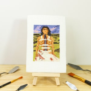 May include: A miniature cross-stitch artwork depicting a figure in a landscape, displayed on a small wooden easel. The artwork features a person in a white dress with a colorful background. Paint knives and a brush surround the artwork.