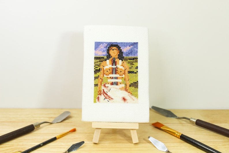 May include: A miniature cross-stitch artwork depicting a figure in a landscape, displayed on a small wooden easel. The artwork features a person in a white dress with a colorful background. Paint knives and a brush surround the artwork.