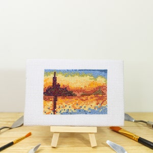 May include: A small, framed cross-stitch landscape depicting a sunset with a dark silhouette of a structure. The artwork is displayed on a miniature wooden easel, surrounded by art tools.