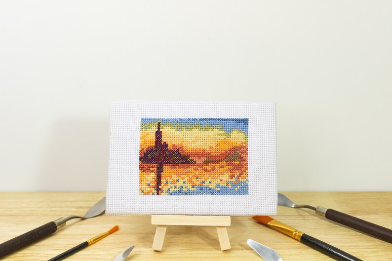 May include: A small, framed cross-stitch landscape depicting a sunset with a dark silhouette of a structure. The artwork is displayed on a miniature wooden easel, surrounded by art tools.