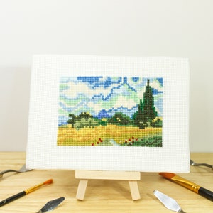 May include: A cross-stitch landscape on a small easel, depicting a vibrant scene with blue skies, green trees, and a yellow field. The artwork is surrounded by art tools, including paintbrushes and palette knives, on a wooden surface.