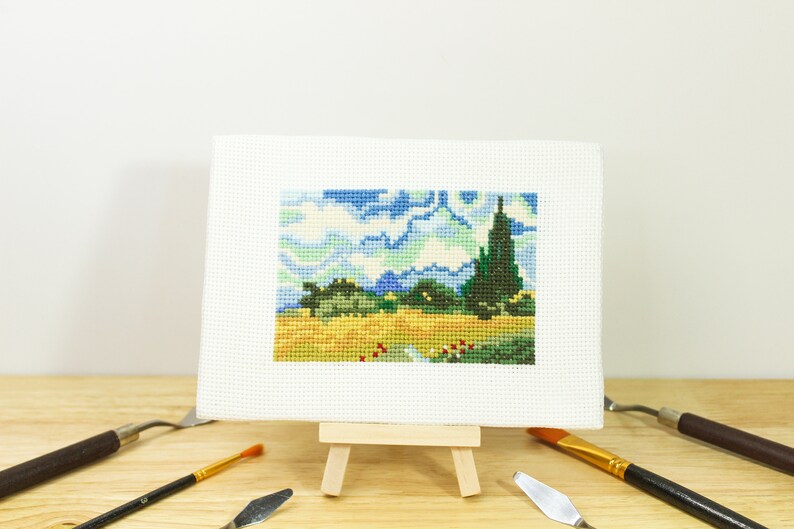 May include: A cross-stitch landscape on a small easel, depicting a vibrant scene with blue skies, green trees, and a yellow field. The artwork is surrounded by art tools, including paintbrushes and palette knives, on a wooden surface.