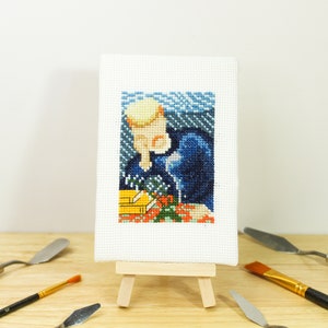 May include: A small, framed cross-stitch artwork depicting a person with a blonde hairstyle, wearing a dark blue outfit, and holding flowers. The artwork is displayed on a miniature wooden easel, surrounded by art tools.