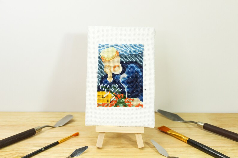 May include: A small, framed cross-stitch artwork depicting a person with a blonde hairstyle, wearing a dark blue outfit, and holding flowers. The artwork is displayed on a miniature wooden easel, surrounded by art tools.