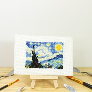 May include: A miniature cross-stitch of Van Gogh's "Starry Night" displayed on a small wooden easel. The artwork features a vibrant blue sky with yellow stars and a crescent moon. Paint knives and a paintbrush surround the artwork.