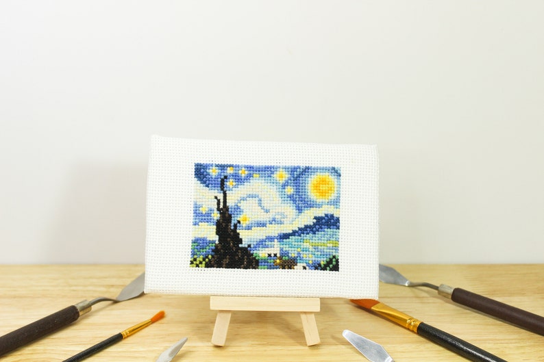May include: A miniature cross-stitch of Van Gogh's "Starry Night" displayed on a small wooden easel. The artwork features a vibrant blue sky with yellow stars and a crescent moon. Paint knives and a paintbrush surround the artwork.