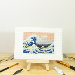 May include: A cross-stitch artwork depicting Hokusai's "The Great Wave off Kanagawa" on a small easel. The wave is rendered in shades of blue, white, and yellow against a peach and gray sky. Art supplies, including paint knives and a brush, are arranged around the artwork.