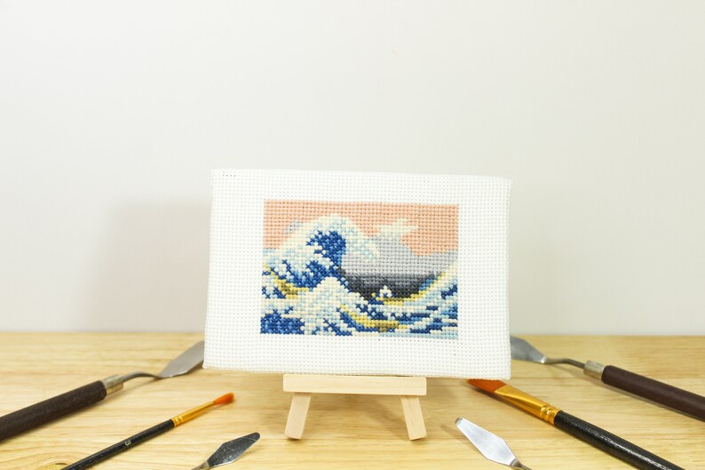 May include: A cross-stitch artwork depicting Hokusai's "The Great Wave off Kanagawa" on a small easel. The wave is rendered in shades of blue, white, and yellow against a peach and gray sky. Art supplies, including paint knives and a brush, are arranged around the artwork.