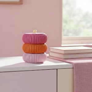 May include: Three stacked, ribbed ceramic containers in pink and orange hues, topped with a small yellow knob. The containers sit on a white surface, with books and a pink runner visible in the background. The scene is set in a room.