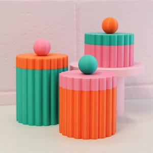 May include: Three cylindrical containers with fluted sides and spherical lids. One container is teal with an orange lid and pink sphere. Another is orange with a pink lid and teal sphere. The third is pink with a teal lid and orange sphere.