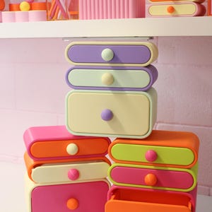 May include: A collection of stackable plastic storage drawers in various pastel and bright colours including purple, light green, yellow, orange, and pink. Each drawer features a single round knob, with some drawers shown open and others closed.
