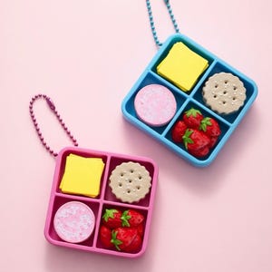 May include: Two miniature lunchbox-style keychains, one blue and one pink, each containing a variety of food items. The blue box has a yellow square, a pink circle, a beige cookie, and red strawberries. The pink box has the same items.