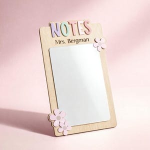 May include: Wooden clipboard with a white writing surface, adorned with pastel-colored letters spelling "NOTES" and the name "Mrs. Bergman." Pink flower accents decorate the corners. The background is a soft pink.