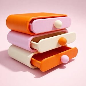 May include: Three-tiered storage drawers in orange, pink, and cream. Each drawer has a contrasting colour pull and a rounded shape. The drawers are stacked on a pink background.