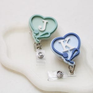 May include: Two heart-shaped badge reels featuring a stethoscope design. One reel is sage green with the white letter J, and the other is navy blue with the white letter K. Each badge reel includes a clear plastic strap and a metal clip attachment.