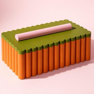 Peut inclure: Une boîte décorative rectangulaire avec un dessus vert festonné et des côtés cylindriques orange. Un objet rose, rectangulaire et arrondi repose sur le dessus. La boîte est posée sur un fond rose.