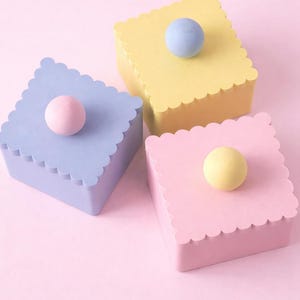 May include: Three pastel-colored square boxes with scalloped edges and a round knob on top. The boxes are light blue, yellow, and pink, set against a pink background. The knobs are in matching colors.