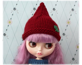 May include: A doll wearing a red knitted hat with a green shamrock embellishment. The doll has pink hair and large blue eyes.
