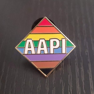 Può includere: Uno spillo AAPI con un design a arcobaleno. Le lettere AAPI sono in bianco su uno sfondo verde, blu, viola, marrone e arancione.