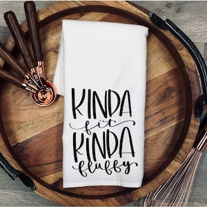 May include: White kitchen towel with black text that reads "Kinda fit kinda fluffy". The towel is on a wooden tray with copper measuring spoons and a copper whisk.