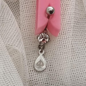 May include: A silver belly button ring with a clear, sparkling gem and a teardrop charm featuring a spade symbol. The ring is held by a pink clip against a white, textured background.