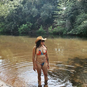 May include: A person wearing a patterned bikini and a cowboy hat stands in a river. The bikini features orange, blue, and white patterns. The person is wearing sunglasses and cowboy boots. The background includes trees and a river.