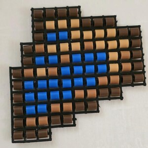 May include: Arrangement of small, cylindrical beads in shades of brown, tan, and blue, set within a black grid. The beads are evenly spaced, creating a patterned design. The overall shape resembles an arrow.