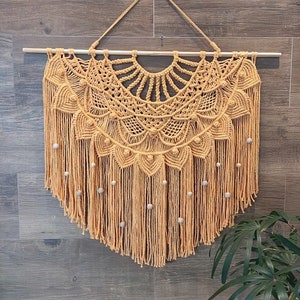 May include: A large, beige macrame wall hanging with a circular design and fringe. The hanging has wooden beads woven into the fringe.