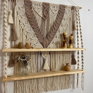 May include: A macrame wall hanging with a brown and cream color pattern, featuring a geometric design. The hanging is attached to two wooden shelves with a rope and tassel detail.