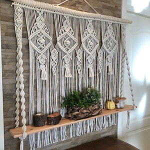 May include: A macrame wall hanging with a wooden dowel rod and a wooden shelf hanging below. The shelf is decorated with a potted plant, candles, and a small pumpkin.