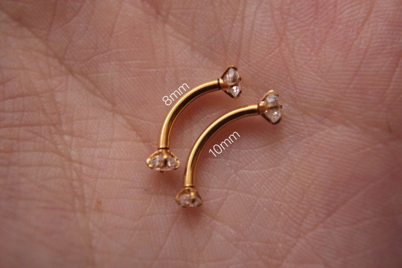 Puede incluir: Dos piezas de joyer&iacute;a corporal de barra curva dorada con gemas de cristal transparente. La pieza superior est&aacute; etiquetada como "8 mm" y la pieza inferior est&aacute; etiquetada como "10 mm".