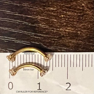 Puede incluir: Dos piezas de joyer&iacute;a corporal de barra curva de color dorado con piedras transparentes en cada extremo. La joyer&iacute;a se mide con una regla, mostrando que la pieza m&aacute;s grande es de 10 mm y la m&aacute;s peque&ntilde;a de 8 mm.