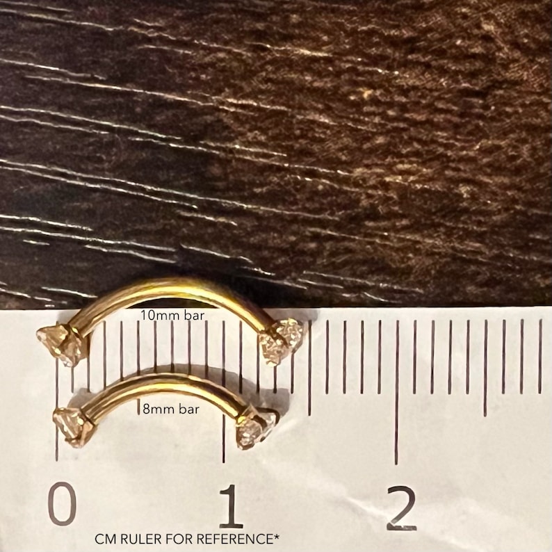 Puede incluir: Dos piezas de joyer&iacute;a corporal de barra curva de color dorado con piedras transparentes en cada extremo. La joyer&iacute;a se mide con una regla, mostrando que la pieza m&aacute;s grande es de 10 mm y la m&aacute;s peque&ntilde;a de 8 mm.