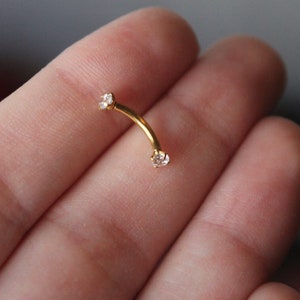 Puede incluir: Joyer&iacute;a de cuerpo dorada con dos piedras preciosas transparentes. La joyer&iacute;a es curva y se sostiene en una mano.