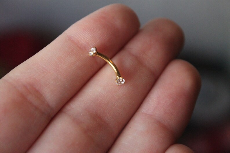 Puede incluir: Joyer&iacute;a de cuerpo dorada con dos piedras preciosas transparentes. La joyer&iacute;a es curva y se sostiene en una mano.