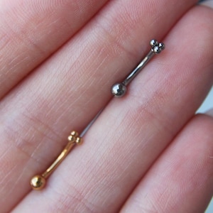 May include: Two curved barbell body jewelry pieces, one in a polished gold tone and one in a polished silver tone. Each barbell features a single smooth ball at one end and a cluster of three small spheres at the opposite end of the curved post.