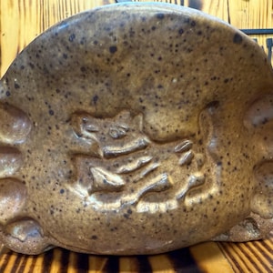 May include: A brown ceramic ashtray with a speckled finish. The ashtray features a raised, stylized design of a creature. The ashtray is resting on a wooden surface with a striped pattern.