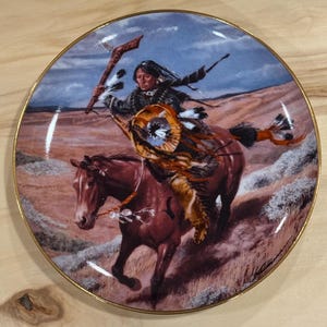 Peut inclure: Assiette décorative avec un guerrier amérindien à cheval, tenant une lance. Le bord de l'assiette est doré. La scène détaillée montre un cheval brun, un guerrier en tenue traditionnelle et un paysage sous un ciel nuageux.