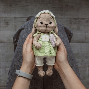 May include: A crocheted bunny toy wearing a green dress with white trim and a white flower. The bunny has brown yarn for its body and white yarn for its face and ears. The bunny is being held by a person's hands.