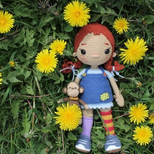 May include: A handmade crochet doll with red pigtails, wearing a blue dress and holding a small monkey. The doll is surrounded by bright yellow dandelions and green grass, creating a whimsical scene. The doll's shoes are blue and the socks are striped.