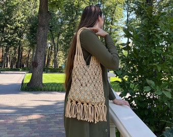May include: A woman wearing a green dress carries a beige crocheted tote bag with fringe. The bag has a net-like design and is worn over the shoulder.