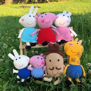 Op de afbeelding: Een verzameling van zeven gehaakte knuffels met verschillende dierfiguren. De groep bevat figuren van roze, wit, geel en bruin garen, gekleed in blauwe, rode en roze outfits, allemaal samen tentoongesteld op een klein houten bankje.