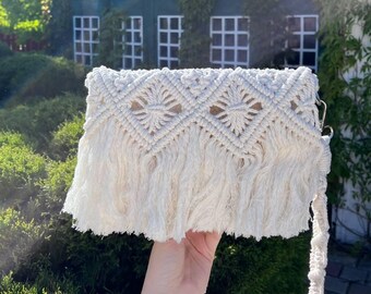 May include: A white crocheted bag with a fringe border and a long strap. The bag has a diamond pattern and is made of cotton.