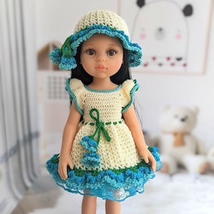 May include: A doll dressed in a cream crochet dress with blue and green details, a matching hat, and blue crochet boots. The dress has a green bow and a ruffled hem. The doll has long black hair and is on a white surface.