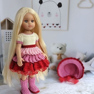 May include: A blonde doll wearing a crocheted dress with pink, red and white layers. The doll is wearing pink crocheted boots and is standing on a white fluffy rug. There is a pink crocheted hat in the background.