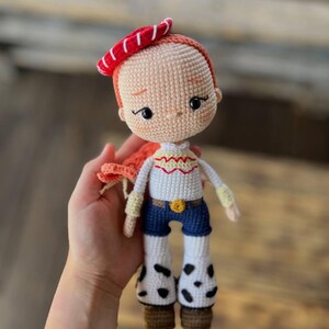 May include: A crocheted doll with red hair, a red and white hat, a white shirt with yellow accents, blue jeans, and black and white cow print boots.