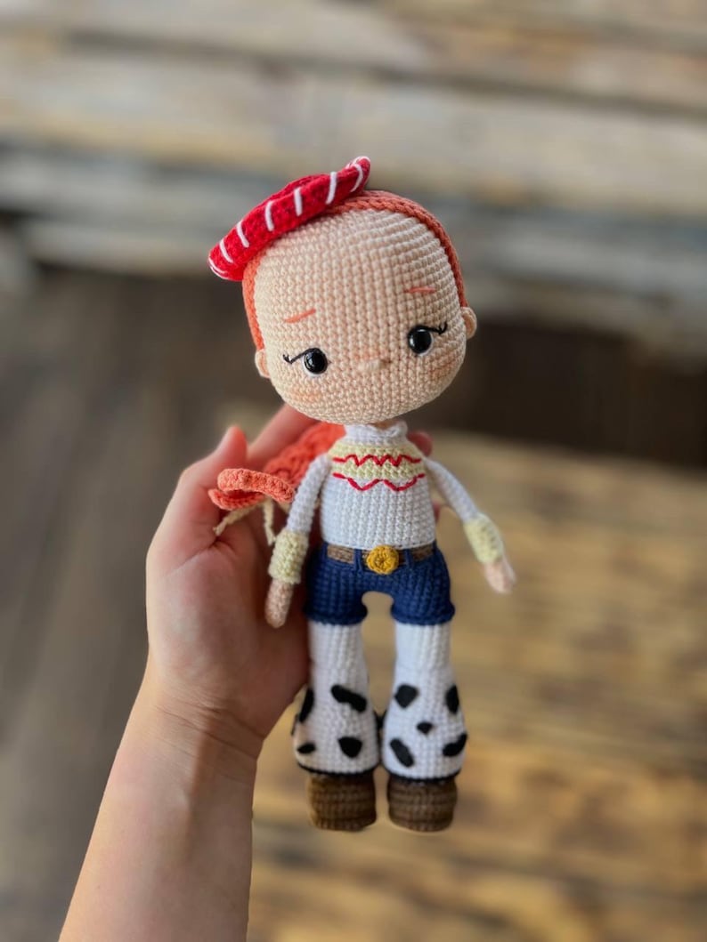 May include: A crocheted doll with red hair, a red and white hat, a white shirt with yellow accents, blue jeans, and black and white cow print boots.