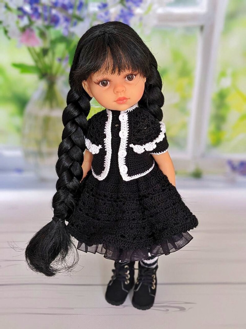 May include: A black and white crocheted dress with a white crocheted collar and sleeves. The doll has long black hair in braids and is wearing black boots.